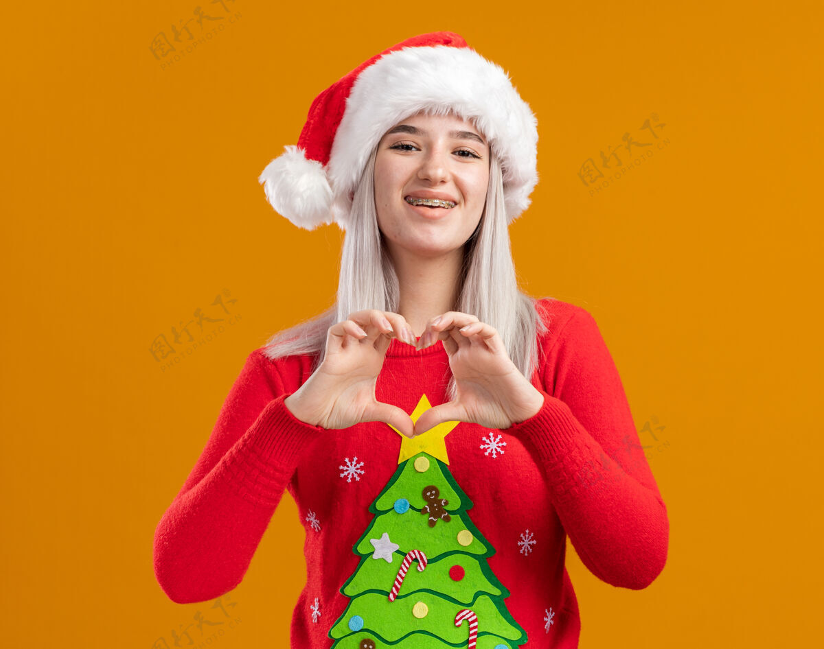 金发穿着圣诞毛衣 戴圣诞帽的金发女郎看着相机 脸上洋溢着幸福的笑容 手指站在橙色背景上做着心形手势圣诞橙色脸