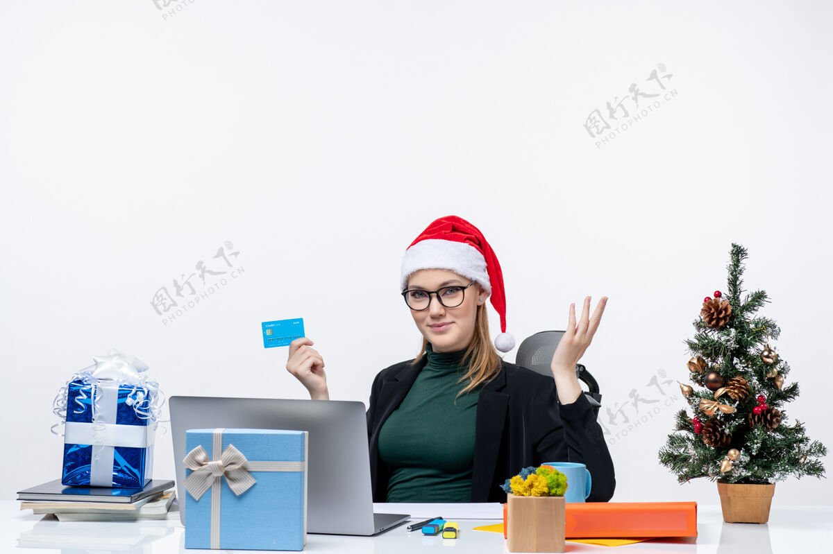 商人戴着圣诞老人帽 戴着眼镜的漂亮女人坐在桌子旁 手里拿着银行卡在办公室问些什么漂亮的桌子电脑
