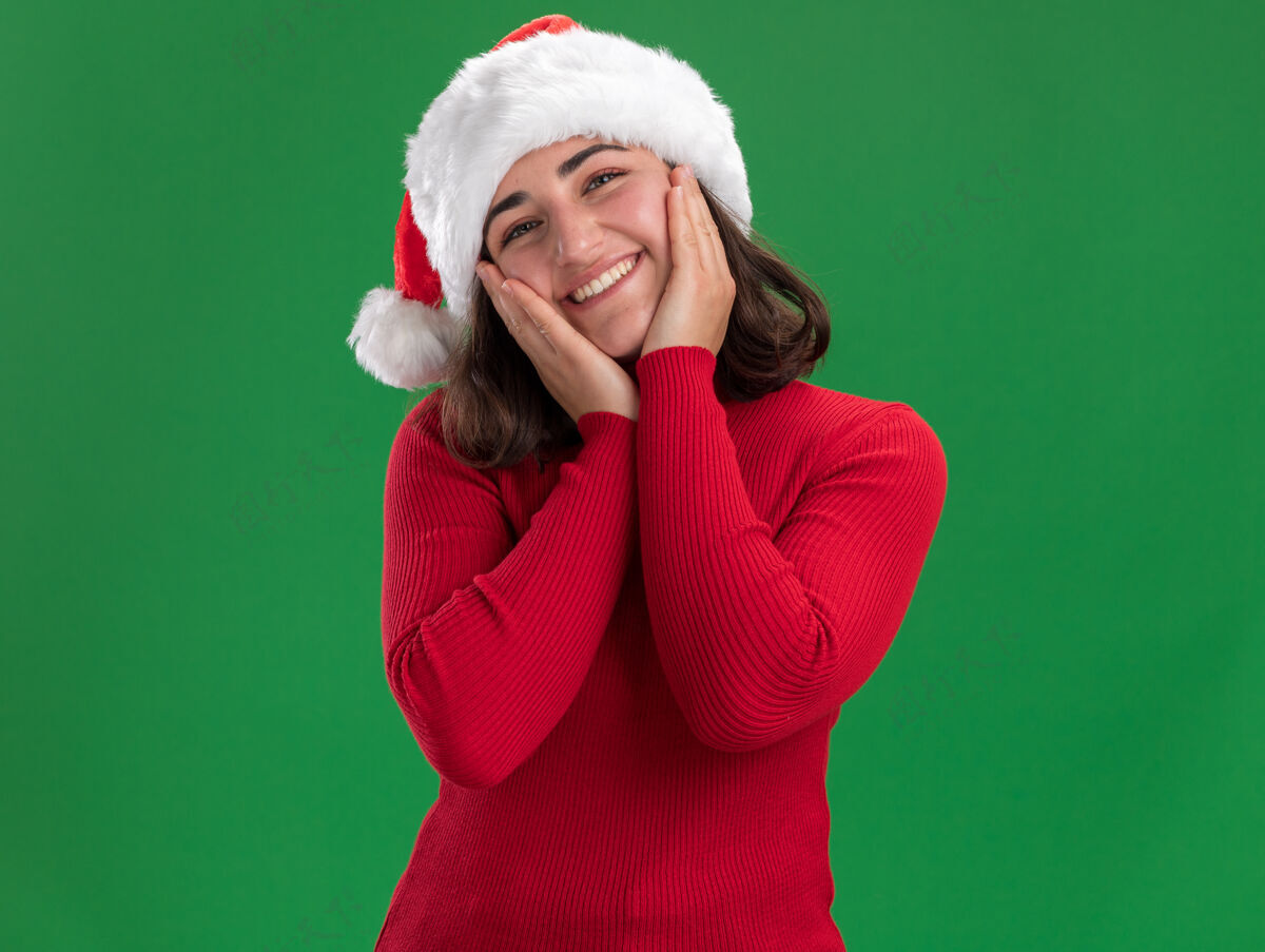 圣诞节穿着红毛衣 戴着圣诞帽的快乐小女孩站在绿色的墙上微笑着帽子宽大站着