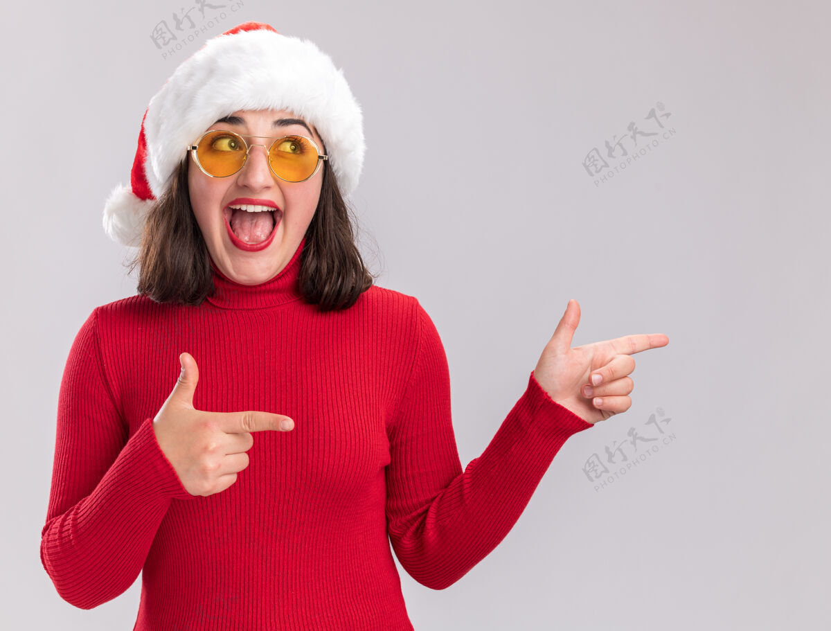 帽子快乐的年轻女孩穿着红色毛衣 戴着圣诞帽 戴着眼镜 一边看一边用食指指着站在白墙上的一边快乐食指毛衣