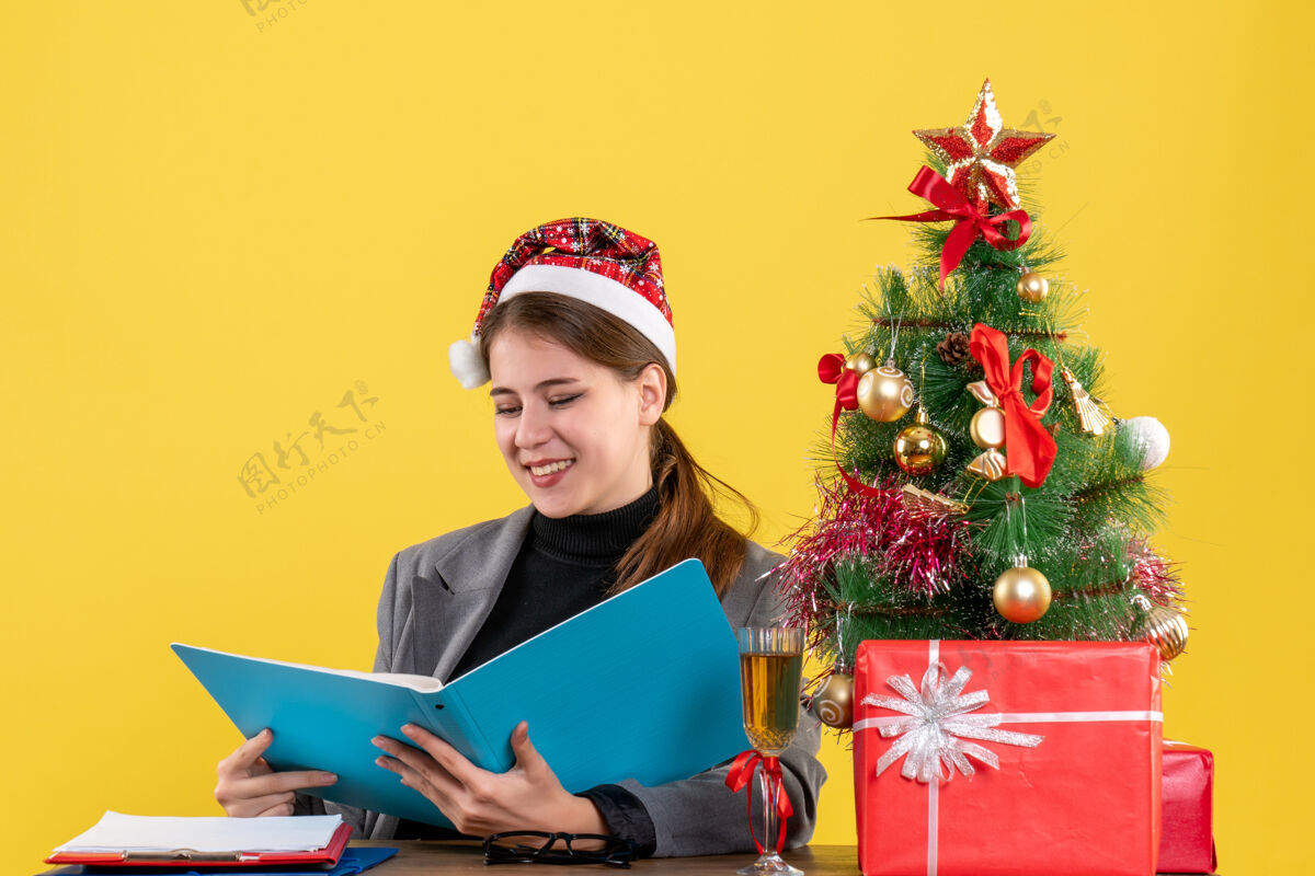 桌子前景笑眯眯的女孩戴着圣诞帽坐在桌旁 看着蓝色文件夹里的文件圣诞树和鸡尾酒礼物文件夹鸡尾酒微笑