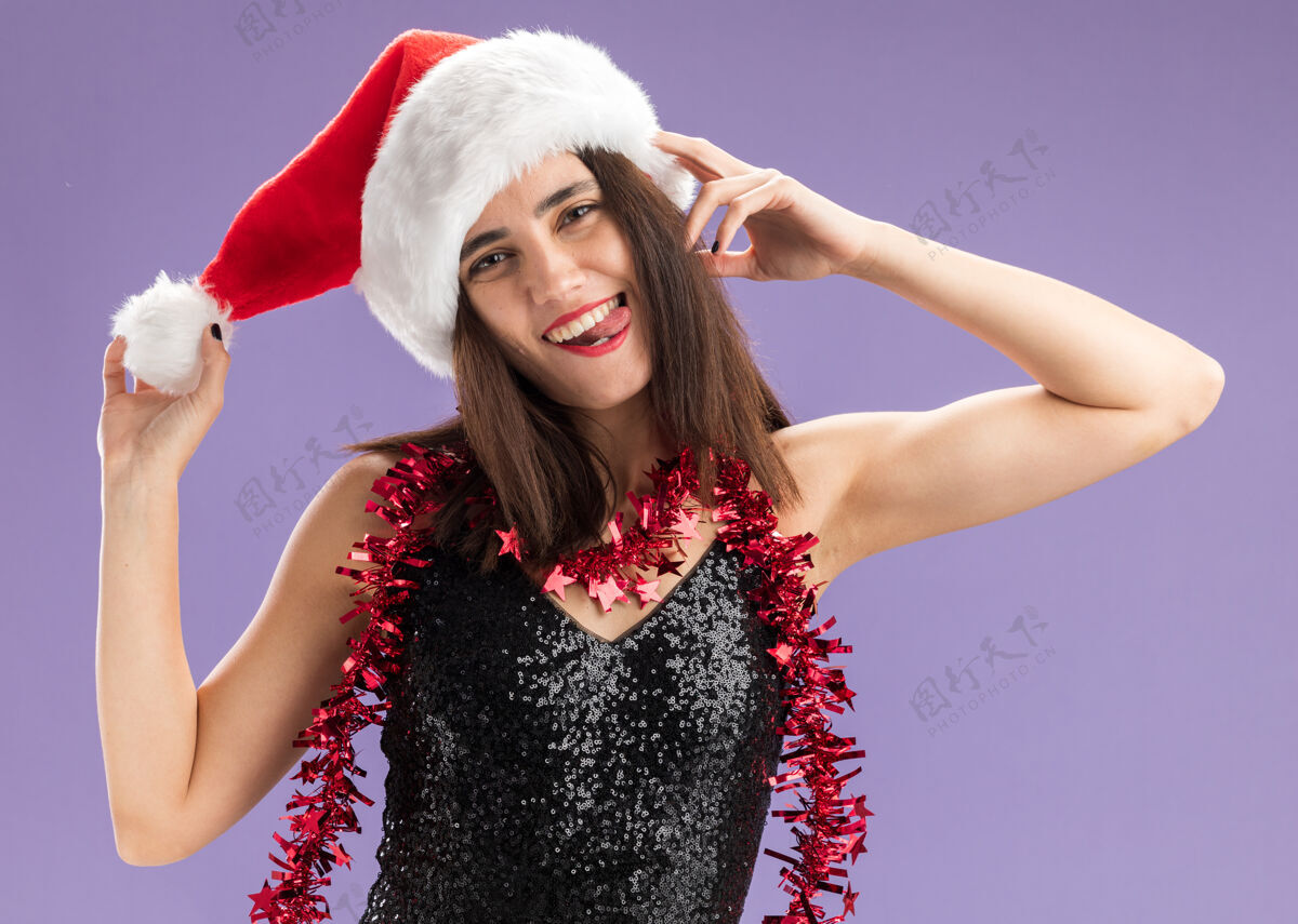 头快乐的歪头年轻漂亮的女孩戴着圣诞帽 脖子上戴着花环 拿着帽子 紫色的墙上露出孤立的舌头脖子帽子快乐
