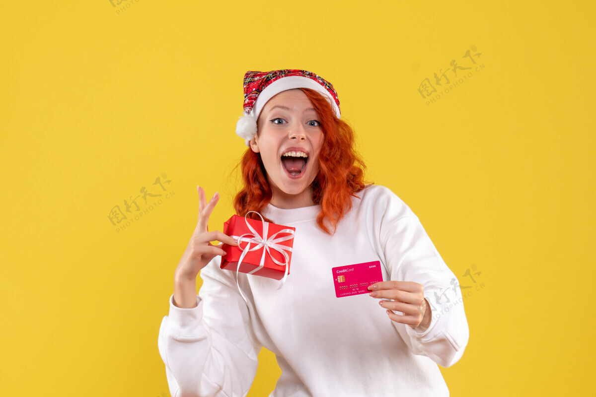 前面圣诞礼物和黄墙上银行卡的年轻女子的正面图圣诞节快乐女人