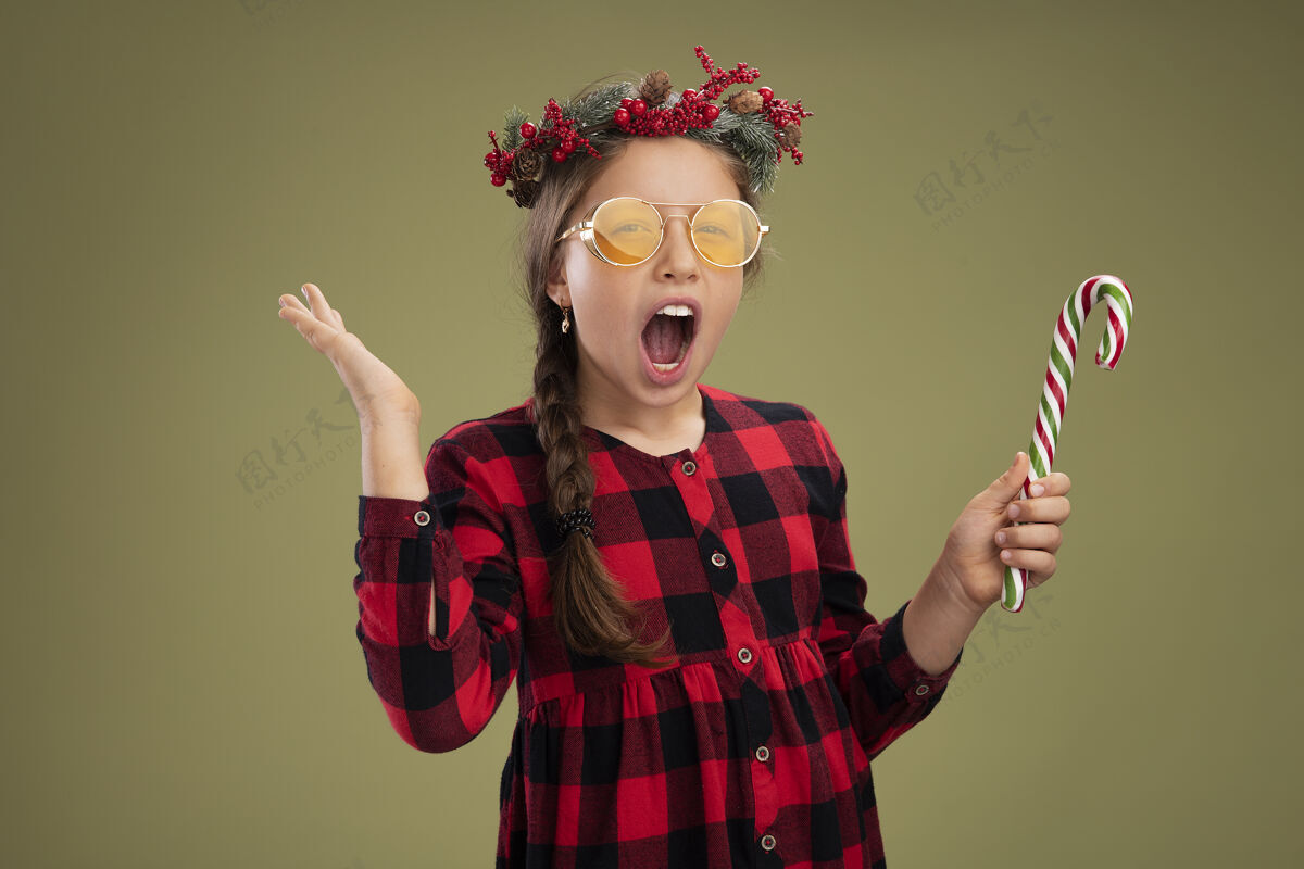 圣诞节小女孩戴着圣诞花圈 穿着格子裙 手持糖果手杖 看着镜头 高兴而兴奋地尖叫着举起手臂站在绿色的背景上抱着衣服手杖