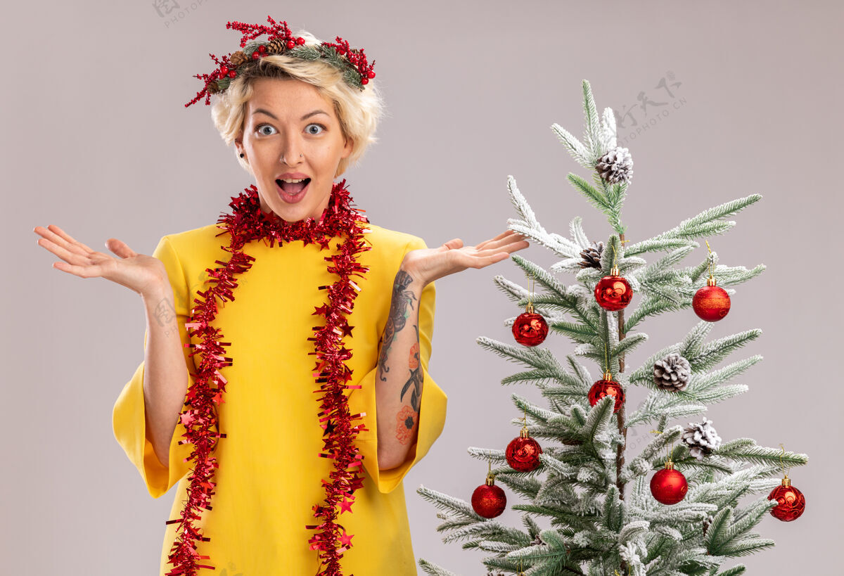 周围年轻的金发女郎戴着圣诞花环 脖子上戴着金箔花环 站在装饰好的圣诞树旁 看着孤零零的双手 孤零零地站在白色的墙上站着花环金发