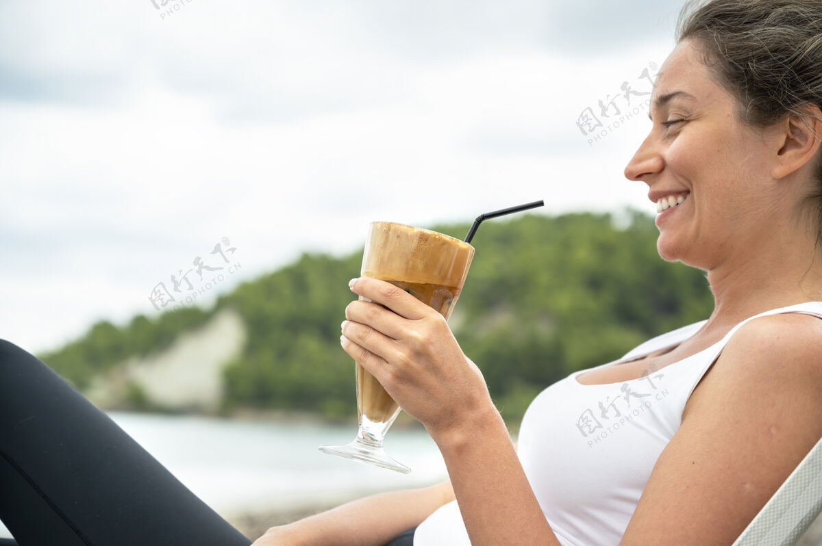 杯子微笑的高加索妇女拿着咖啡饮料在沙滩上与泡沫和喝稻草与山沉思热周到