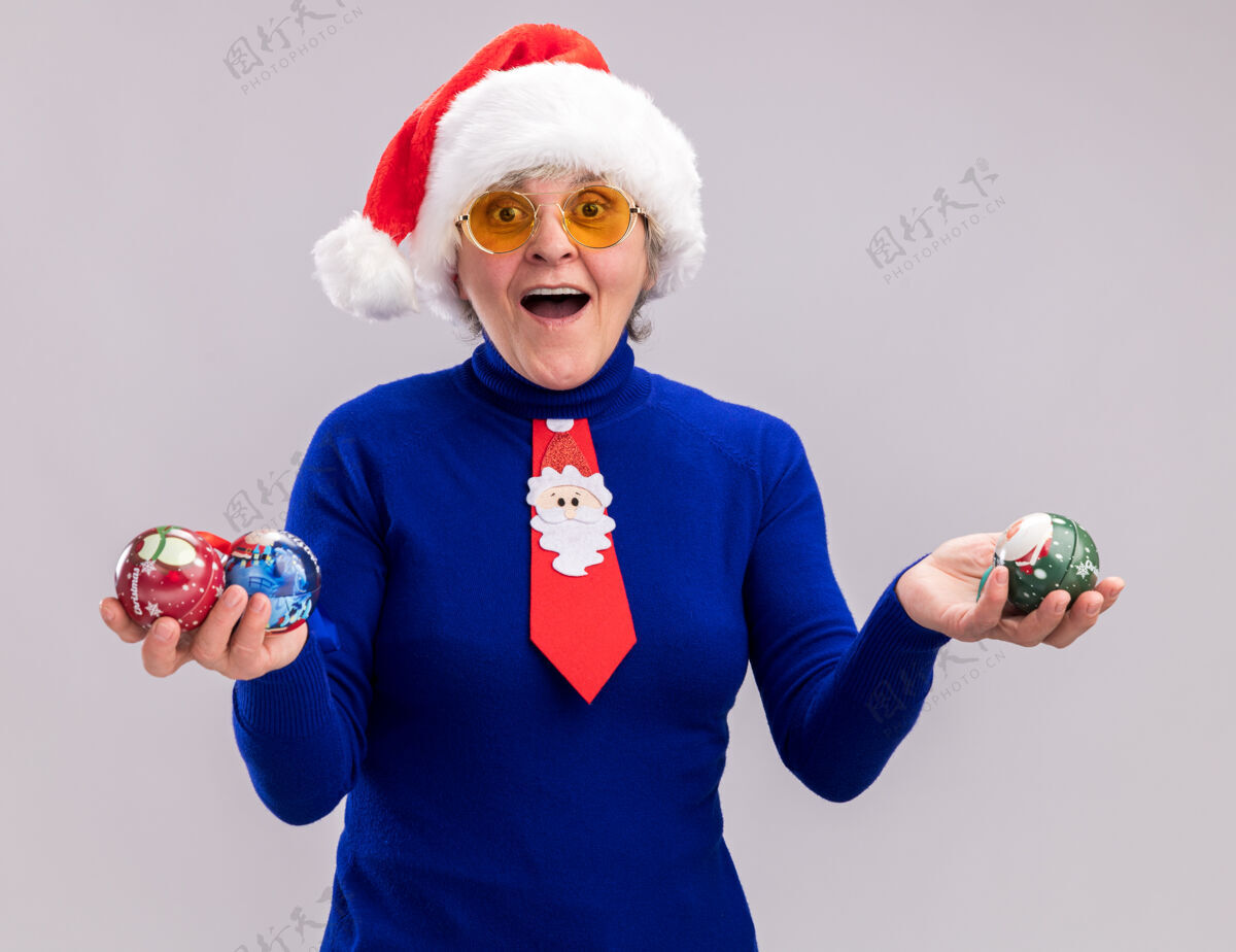 快乐兴奋的老太婆戴着太阳眼镜 戴着圣诞帽 打着圣诞领带 手里拿着玻璃球饰品 在白色背景上与复制空间隔离开来眼镜新年年