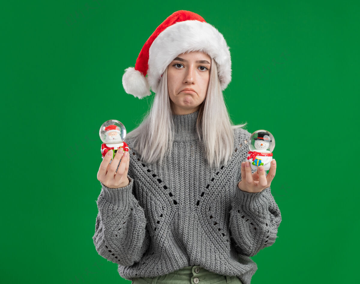 快乐年轻的金发女郎穿着冬季毛衣 戴着圣诞帽 手里拿着圣诞玩具雪球 神情悲伤地看着相机 噘着嘴唇站在绿色的背景上抱着圣诞快乐Pursing