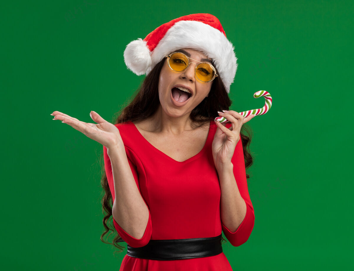 漂亮令人印象深刻的年轻漂亮女孩戴着圣诞帽和眼镜拿着圣诞糖果手杖看着相机显示空手孤立在绿色的背景抱着空帽子