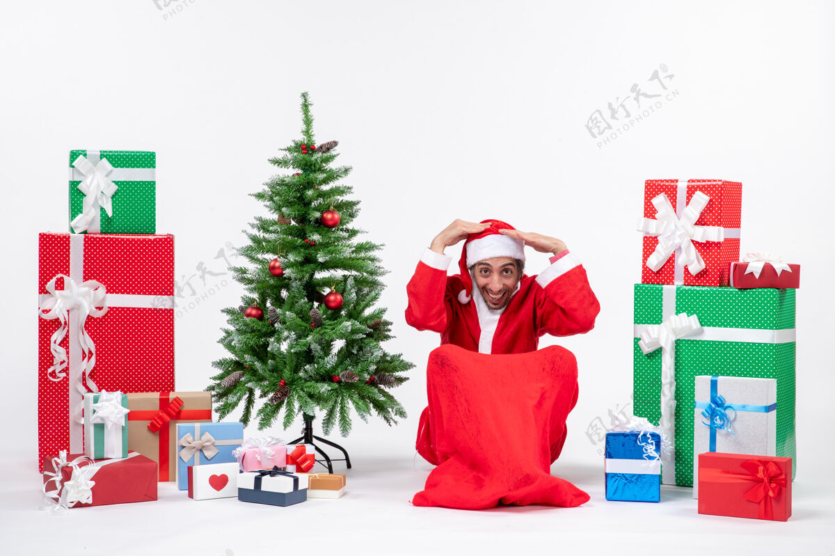 帽子年轻人打扮成圣诞老人 拿着礼物和装饰好的圣诞树坐在地上双手合十快乐圣诞老人手