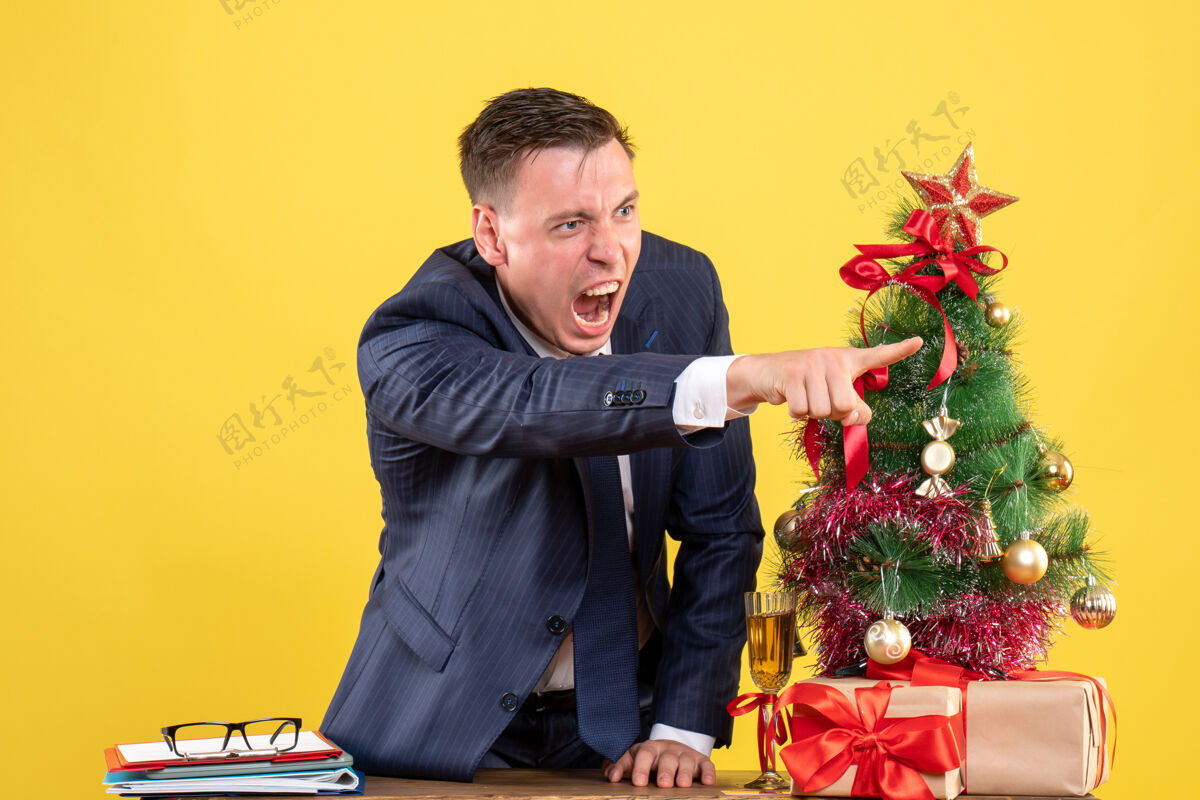 某人愤怒的男人在圣诞树旁的桌子后面对某人喊叫 黄色的墙上有礼物圣诞男性人