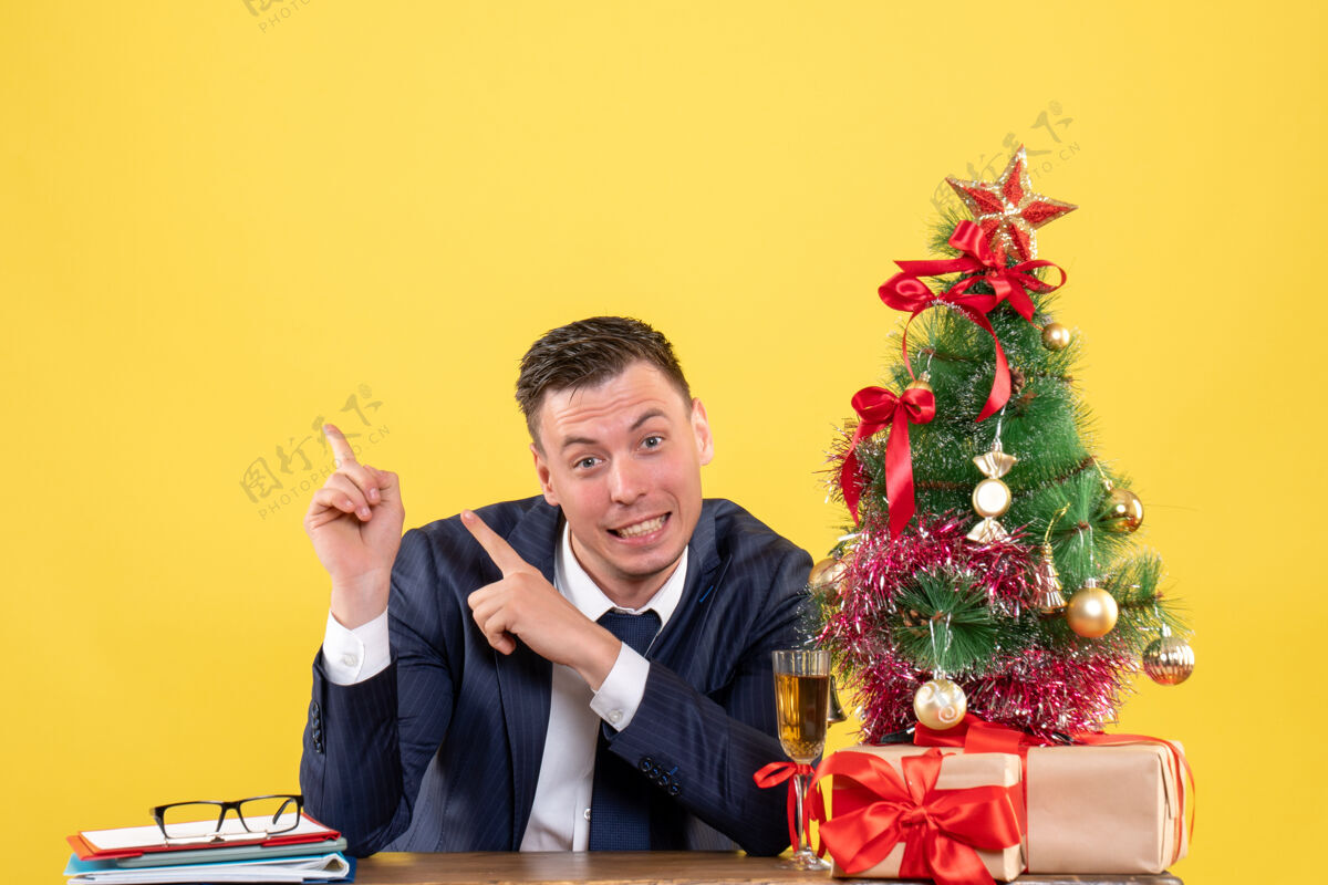 西装正面图微笑的男人指着坐在圣诞树旁桌子上的东西和黄色的礼物办公室男黄色