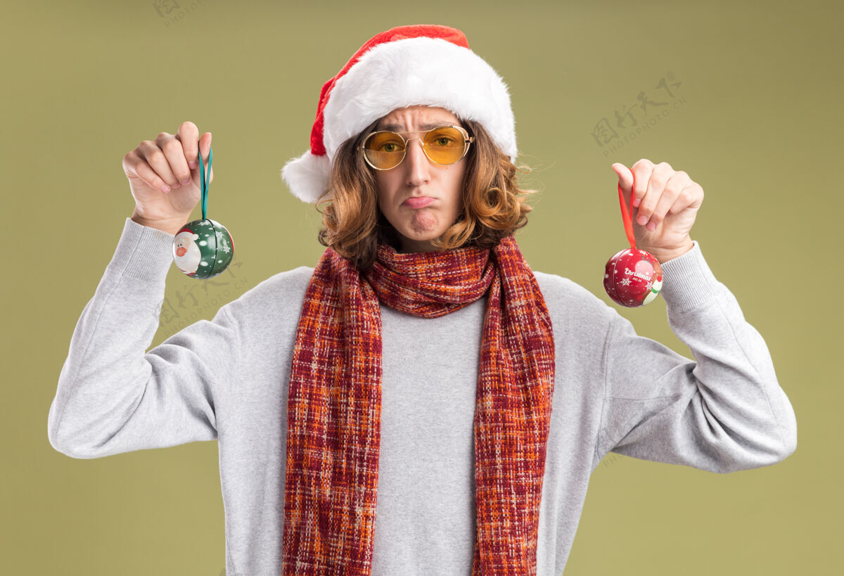 相机沮丧的年轻人戴着圣诞老人的帽子 戴着黄色的眼镜 脖子上围着暖和的围巾 手里拿着圣诞球 看着相机 脸上带着悲伤的表情 噘着嘴唇站在绿色的背景上抱着站着表情