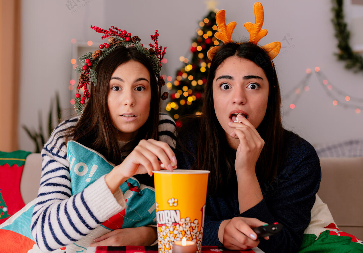 坐着带着冬青花环和驯鹿头带的年轻女孩们吃着爆米花 坐在扶手椅上看电视 在家里享受圣诞节的快乐电视快乐冬青