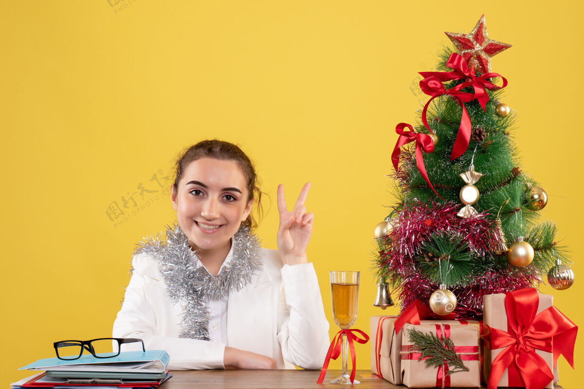 背景正面图：女医生坐在黄色背景的桌子后面 还有圣诞树和礼品盒插花人女医生