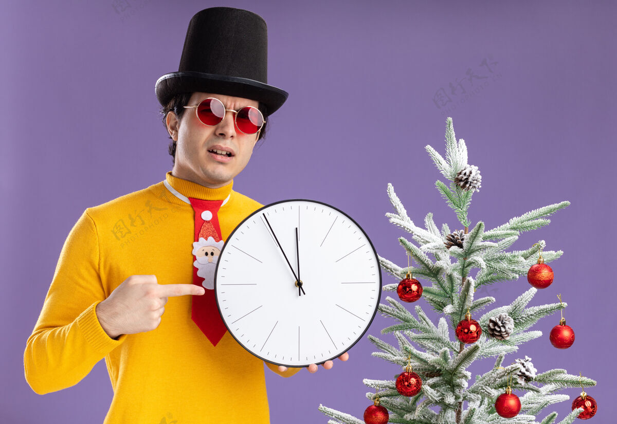 手指身穿黄色高领毛衣 戴着黑帽子 戴着眼镜的年轻人拿着挂钟 用食指指着挂钟 表情严肃地站在紫色墙上的圣诞树旁食指高领毛衣圣诞节