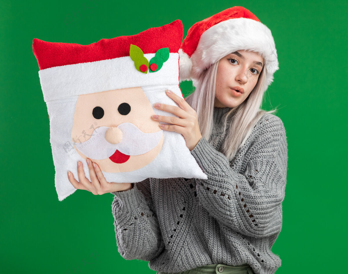 站着年轻的金发女郎穿着冬衣 戴着圣诞帽 抱着圣诞枕头 站在绿色的背景下 自信地看着镜头毛衣抱着年轻