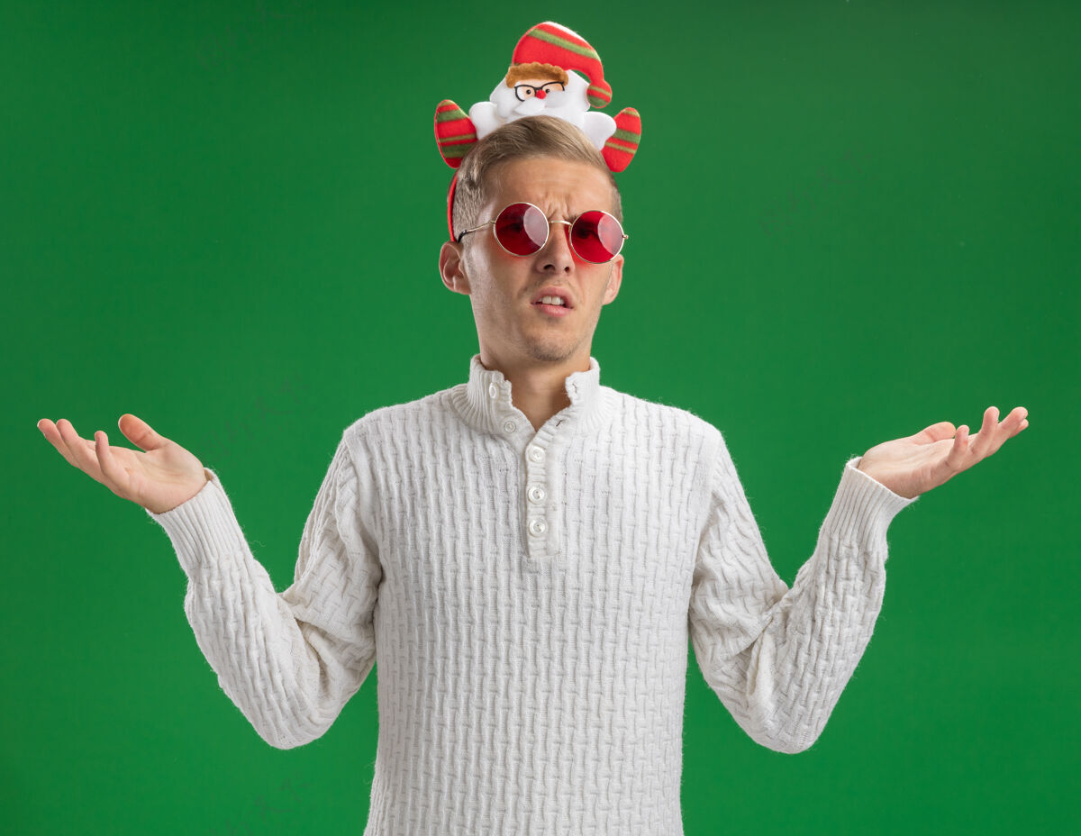 空虚无知的年轻帅哥戴着圣诞老人的头带 戴着眼镜 两手空空的 孤零零地站在绿色的墙上穿着头带不知所措
