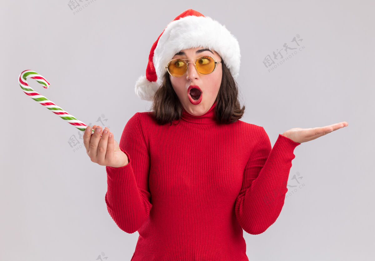 年轻穿着红色毛衣 戴着圣诞帽 戴着眼镜 拿着糖果手杖的年轻女孩一边看着 一边惊讶 一边困惑地举起双臂站在白色的墙上帽子手杖毛衣