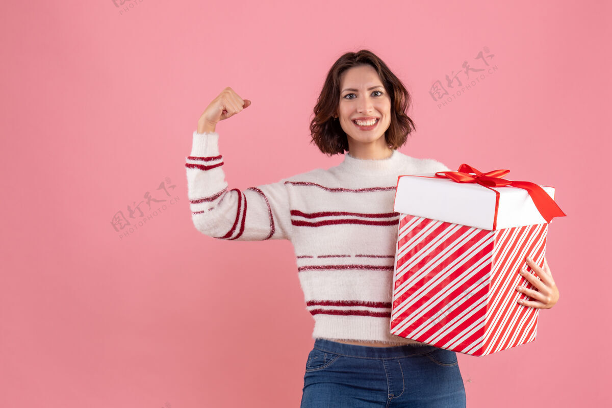 年轻带着圣诞礼物的年轻女子在粉色墙上微笑的正面图圣诞节年轻女性节日