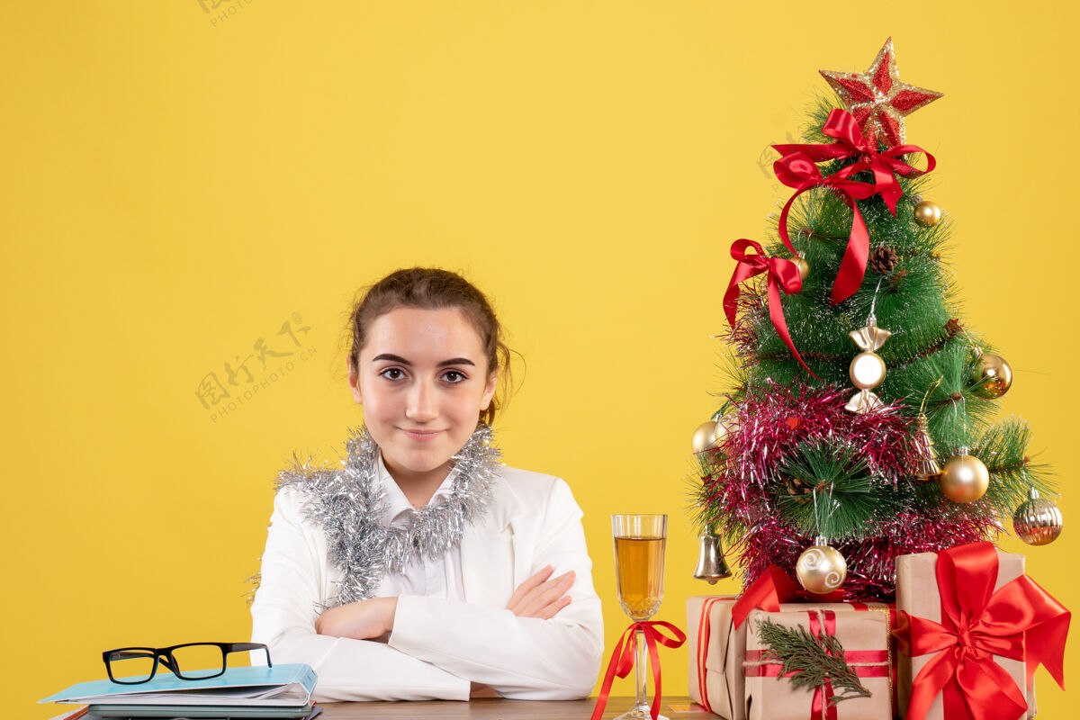 插花正面图：女医生坐在黄色背景的桌子后面 还有圣诞树和礼品盒圣诞节快乐坐着