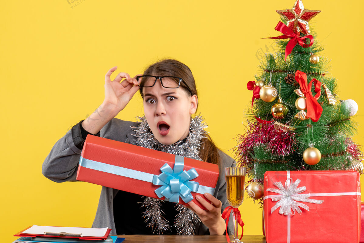 欢快前视图好奇的女孩坐在桌边摘下眼镜圣诞树和鸡尾酒礼物树眼镜坐着