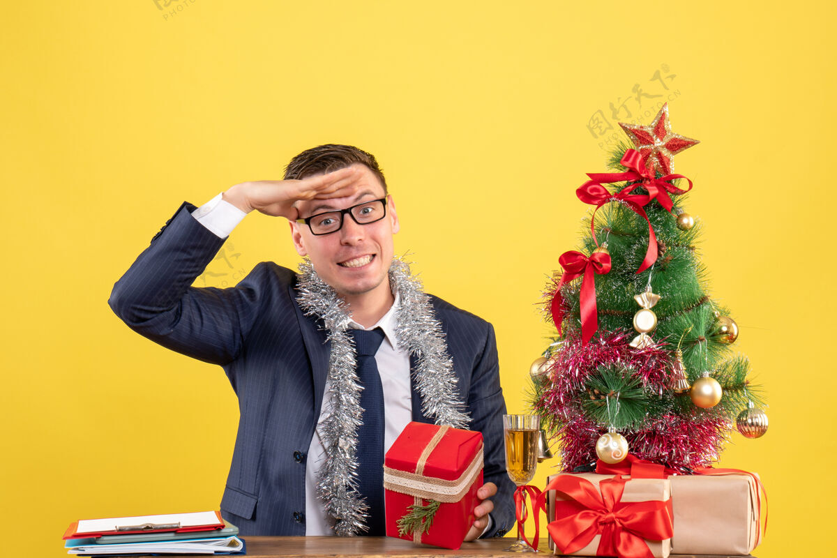 新郎正面图：快乐的男人把手放在额头上 坐在圣诞树旁的桌子旁 黄色的礼物男人礼物黄色