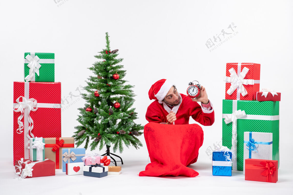 看年轻人坐在地上 看着圣诞树旁的礼物和装饰的时钟庆祝圣诞节年轻人装饰礼物