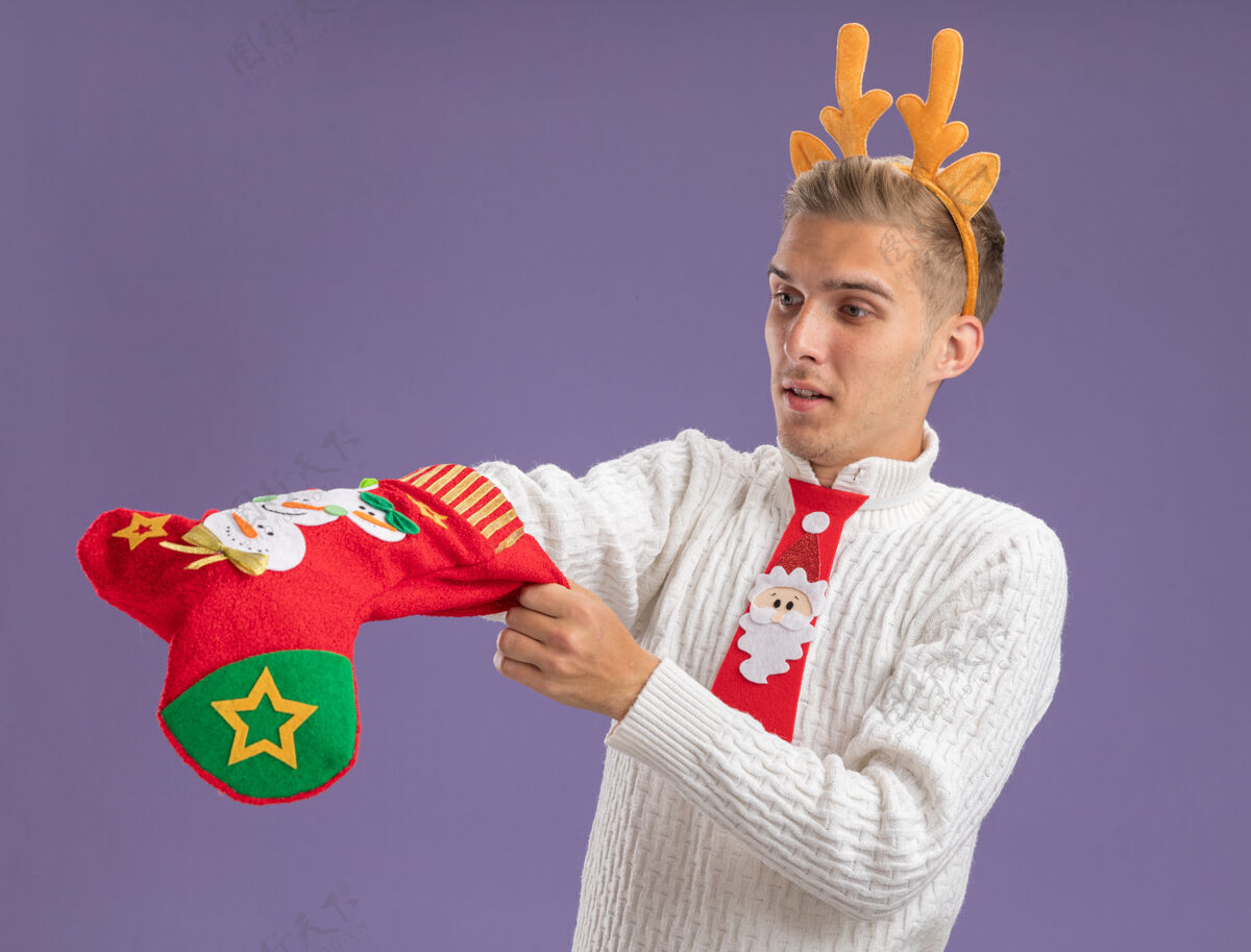 驯鹿好奇的年轻帅哥戴着驯鹿的鹿角头带 打着圣诞老人的领带 手里拿着圣诞长袜 看着里面的手孤立在紫色的墙上戴领带圣诞节