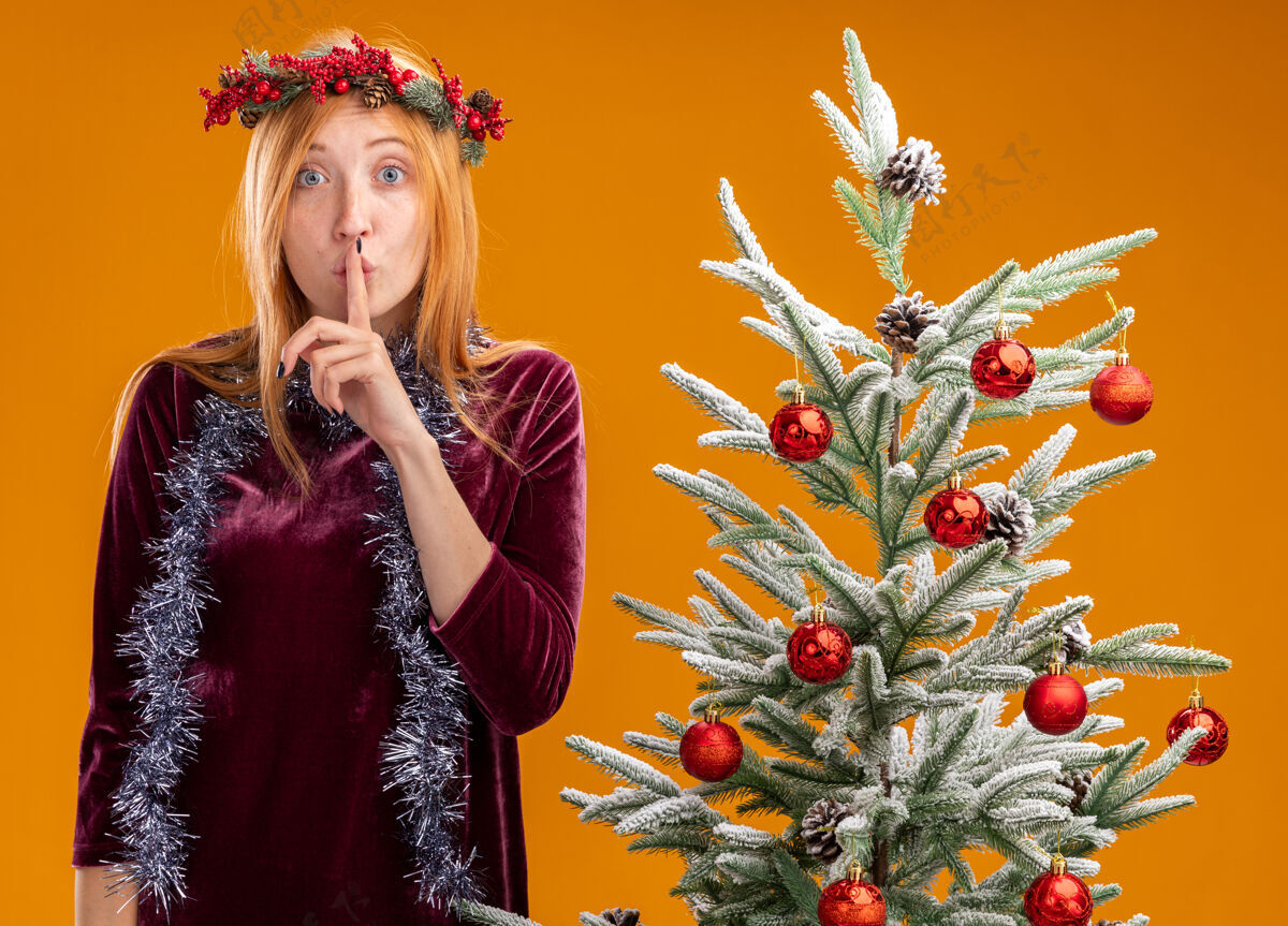 树站在圣诞树旁的年轻漂亮的女孩 穿着红色的裙子 脖子上戴着花环 在橙色的背景下显示出沉默的姿态手势脖子圣诞节
