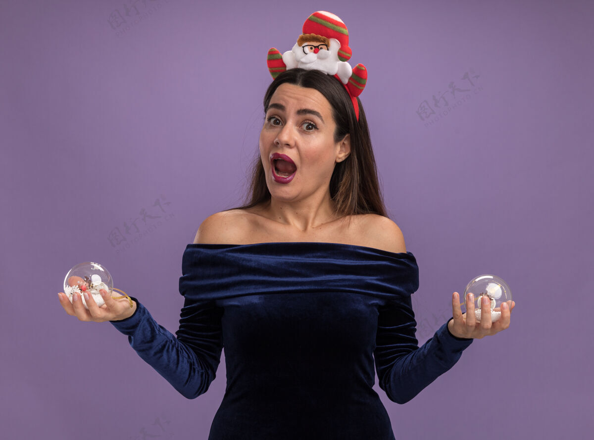 惊喜惊讶的年轻漂亮女孩穿着蓝色连衣裙 戴着圣诞发箍 手里拿着紫色背景上孤立的圣诞球女孩篮球圣诞节