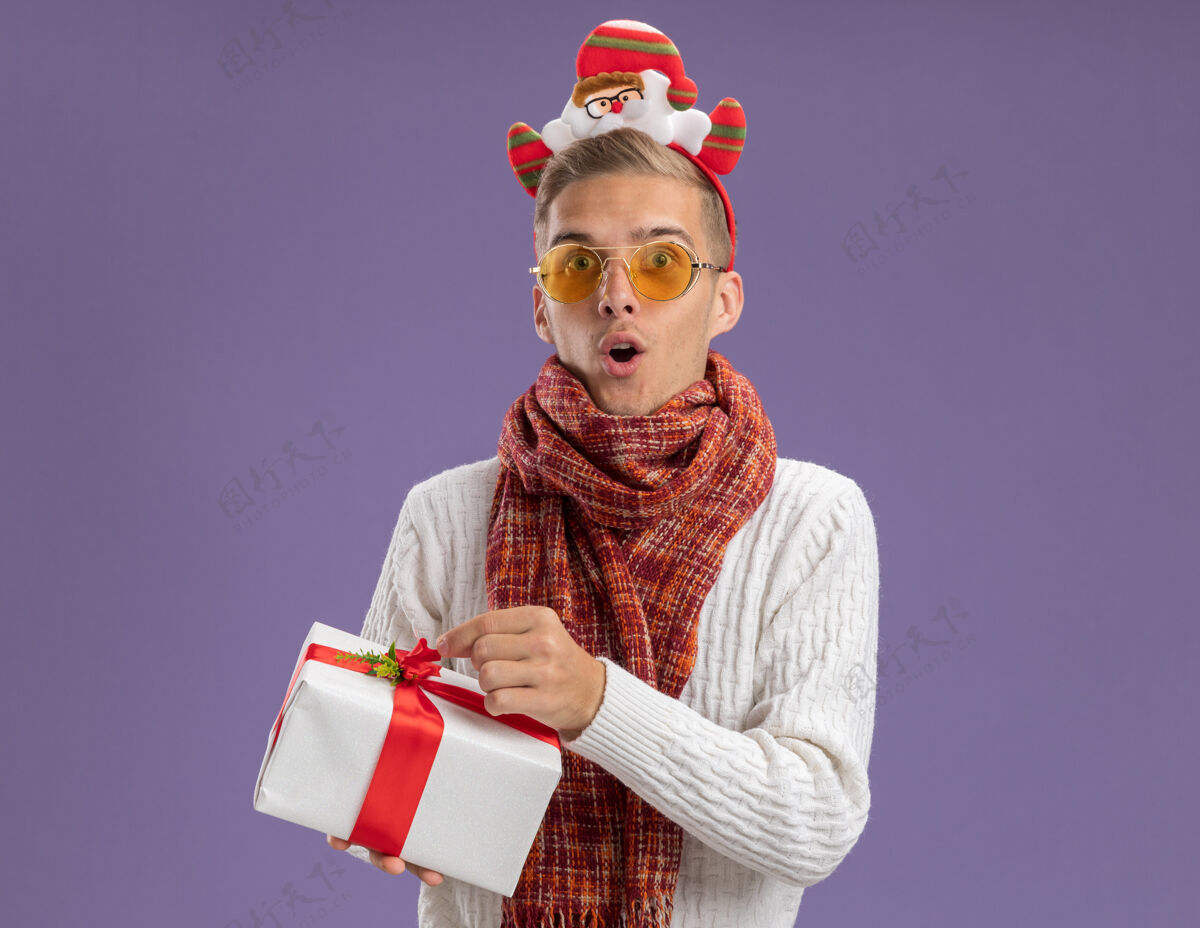 家伙惊讶的年轻帅哥戴着圣诞老人的头带和围巾拿着礼包看着相机隔离在紫色背景上的复制空间围巾礼物举行