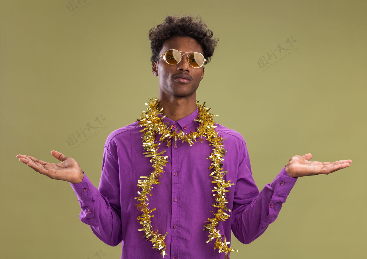 显示年轻的非裔美国人戴着眼镜 脖子上戴着金箔花环 两手空空的 孤零零地站在橄榄绿的墙上圣诞节年轻男人