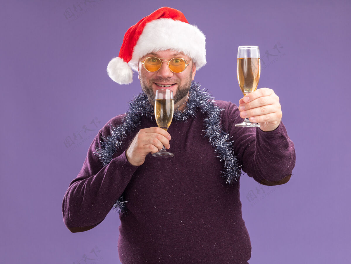 圣诞老人微笑的中年男子戴着圣诞帽 脖子上戴着金箔花环 戴着一副眼镜 手里拿着两杯香槟 一杯孤立地放在紫色的墙上戴着帽子男人