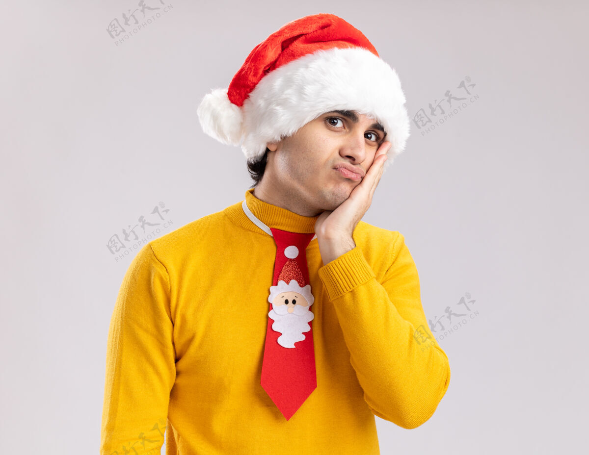 领带一个穿着黄色高领毛衣 戴着圣诞帽 打着滑稽领带的年轻人站在白色背景下 看着镜头 又累又无聊站着帽子疲倦