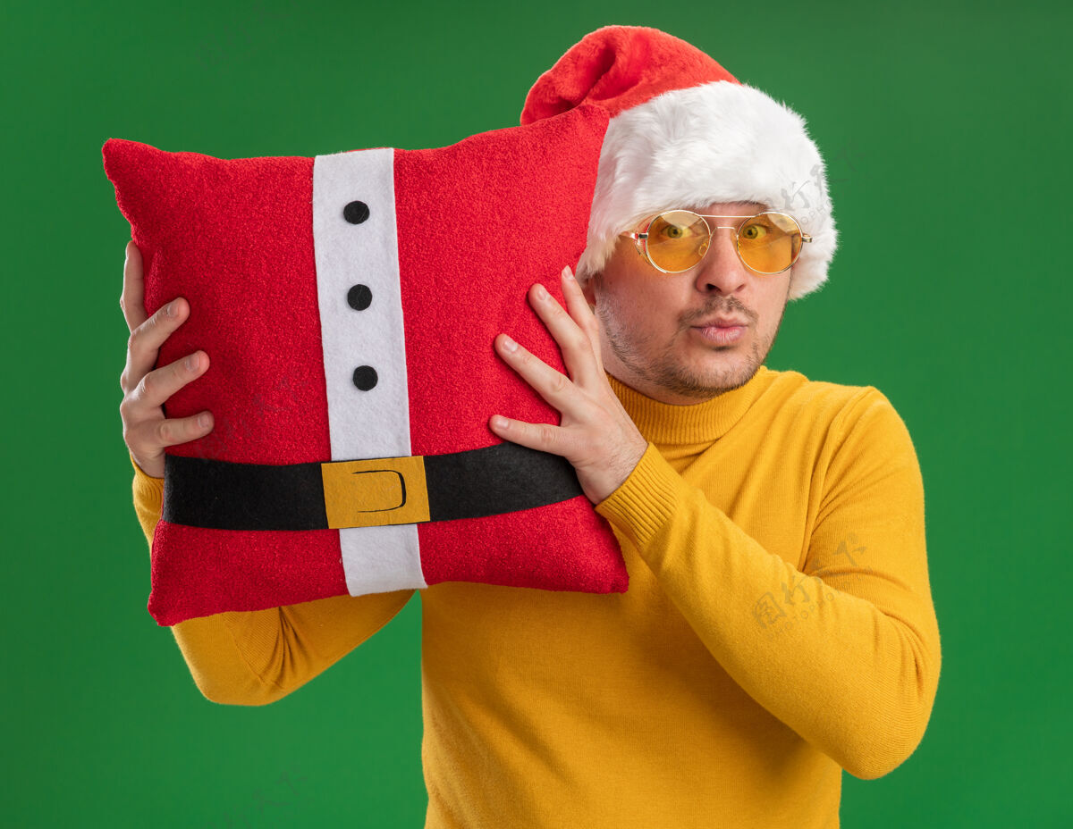 眼镜一个穿着黄色高领毛衣 戴着圣诞帽 戴着眼镜 拿着红色滑稽枕头的年轻人站在绿色背景下惊讶地看着摄像机惊讶圣诞老人帽子