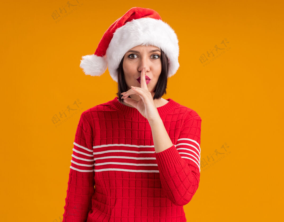 印象深刻令人印象深刻的年轻女孩戴着圣诞帽看着相机做沉默的手势 在橙色的背景与复制空间隔离帽子圣诞老人年轻