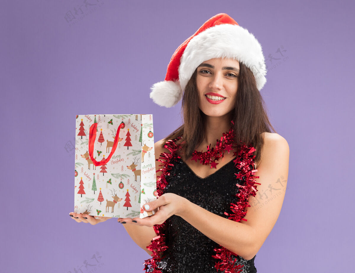 年轻微笑着的年轻漂亮女孩戴着圣诞帽 脖子上戴着花环 手里拿着紫色背景上孤立的礼品袋圣诞节女孩帽子