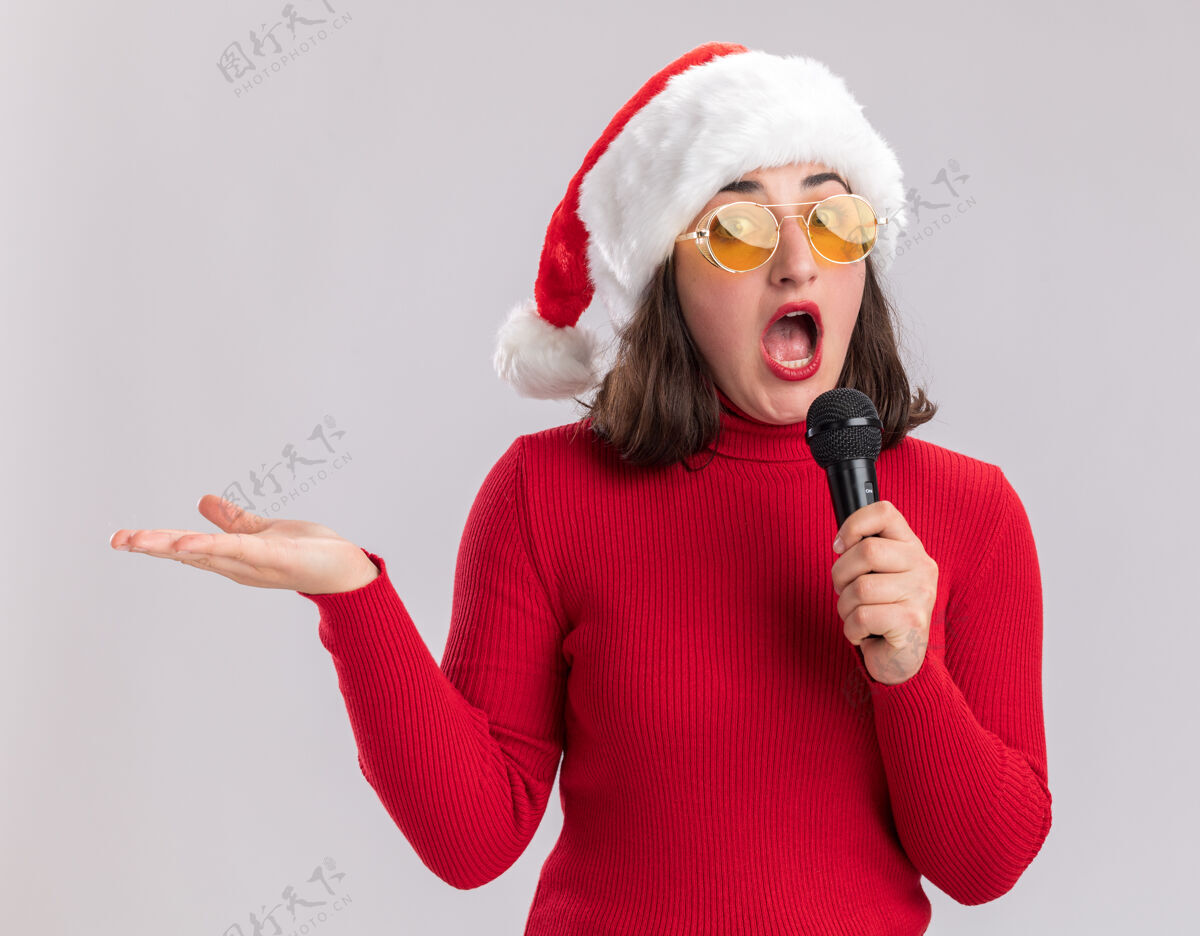眼镜穿着红色毛衣 戴着圣诞帽 戴着眼镜 拿着麦克风 惊讶地看着镜头 伸出手臂站在白色背景上毛衣麦克风女孩