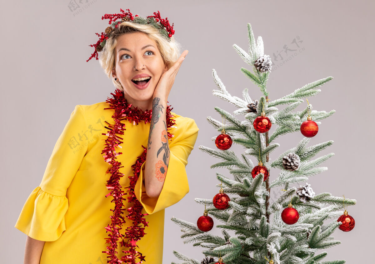触摸年轻的金发女郎戴着圣诞花环 脖子上戴着金箔花环 站在装饰好的圣诞树旁 抚摸着脸 看着隔离在白色墙上的一面印象深刻站着头