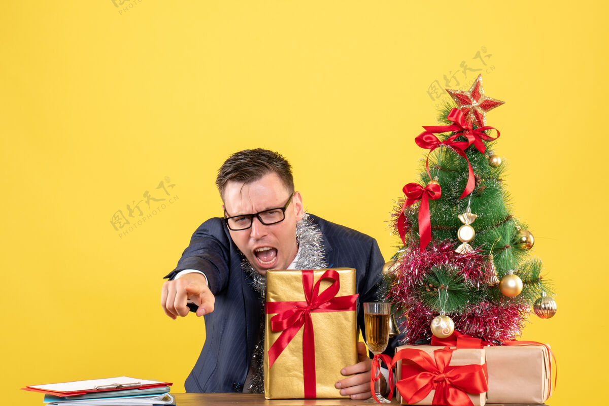 套装前视图愤怒的男子指着摄像机坐在圣诞树附近的桌子和黄色墙上的礼物快乐人指着