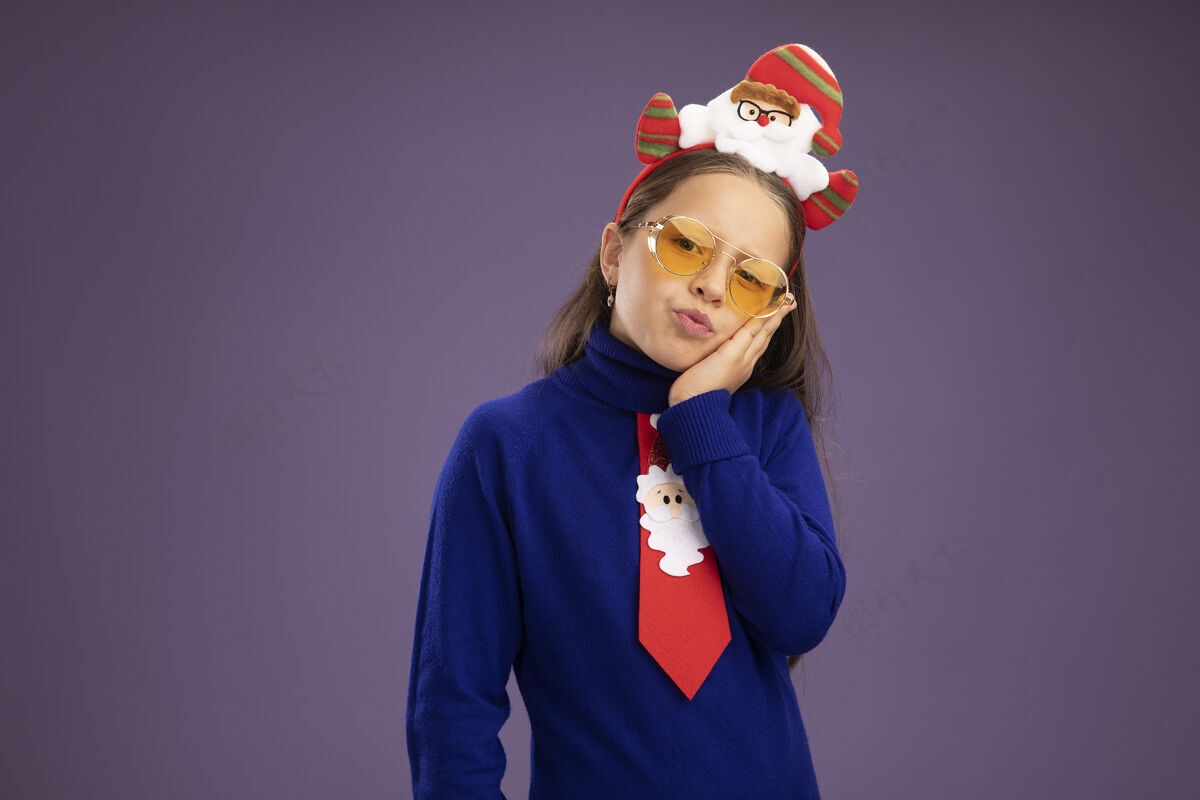 有趣小女孩穿着蓝色高领毛衣 打着红色领带 头上戴着有趣的圣诞戒指 站在紫色的背景下 看着镜头 又累又无聊边缘头累