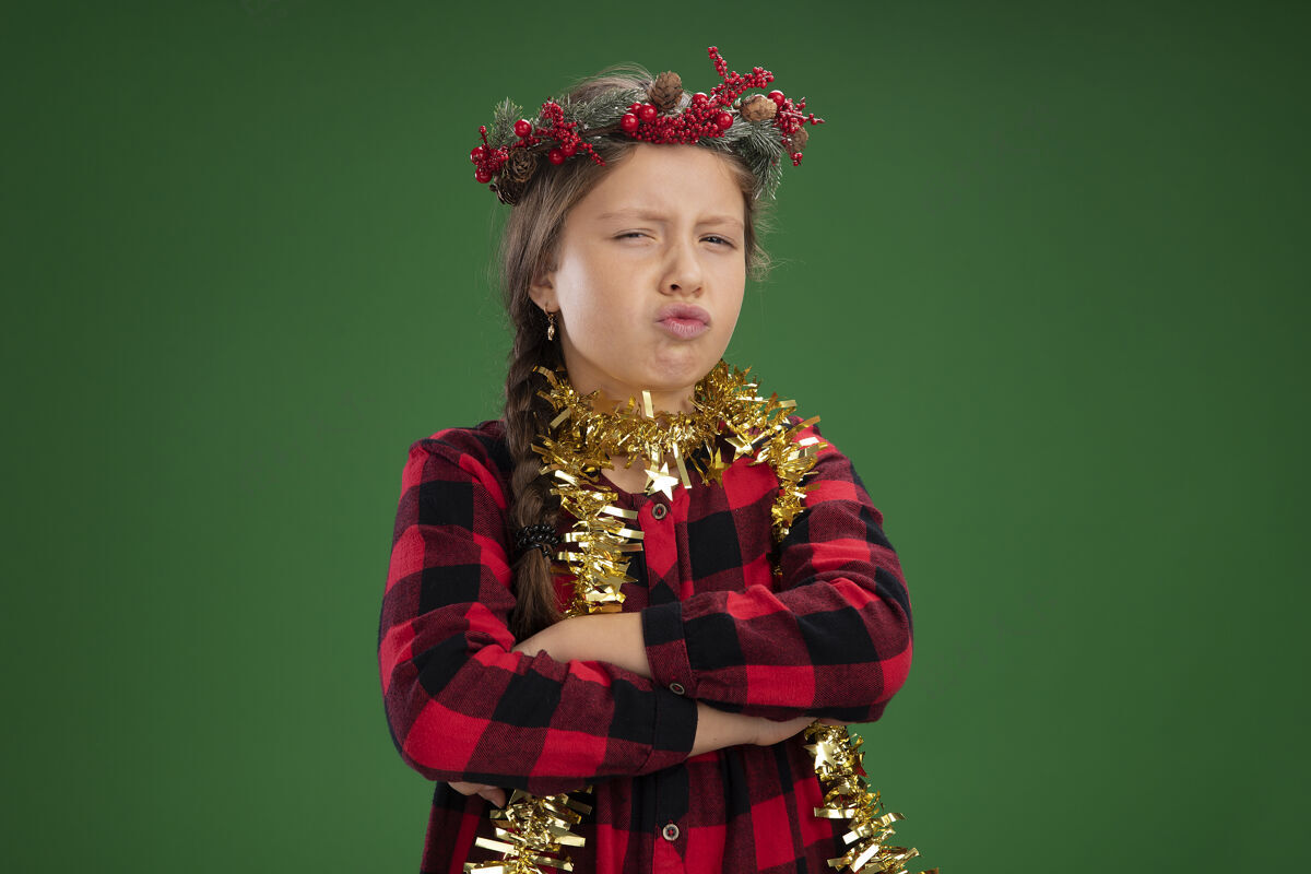 胳膊愤愤不平的小女孩戴着圣诞花环 身穿格子裙 脖子上戴着金属丝 皱着眉头 双臂交叉着站在绿色背景上看着摄像机衣服周围女孩