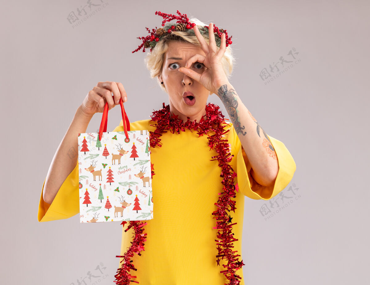 花环令人印象深刻的年轻金发女子头戴圣诞花环 脖子上戴着金属丝花环 手里拿着圣诞礼品袋 看起来像在做一个孤立在白墙上的手势看头手势