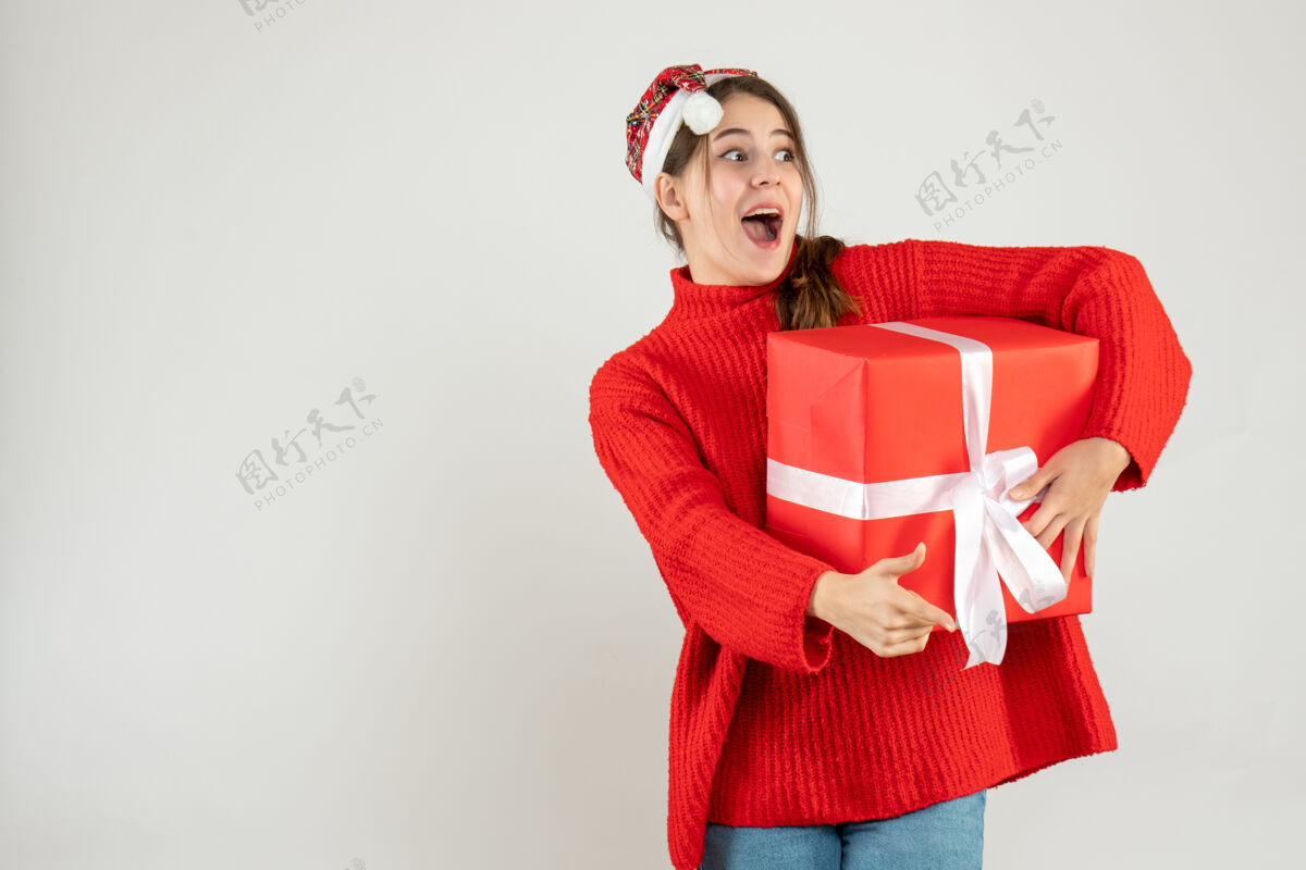 可爱戴着圣诞帽的快乐女孩手指着拿着礼物的东西漂亮指着帽子