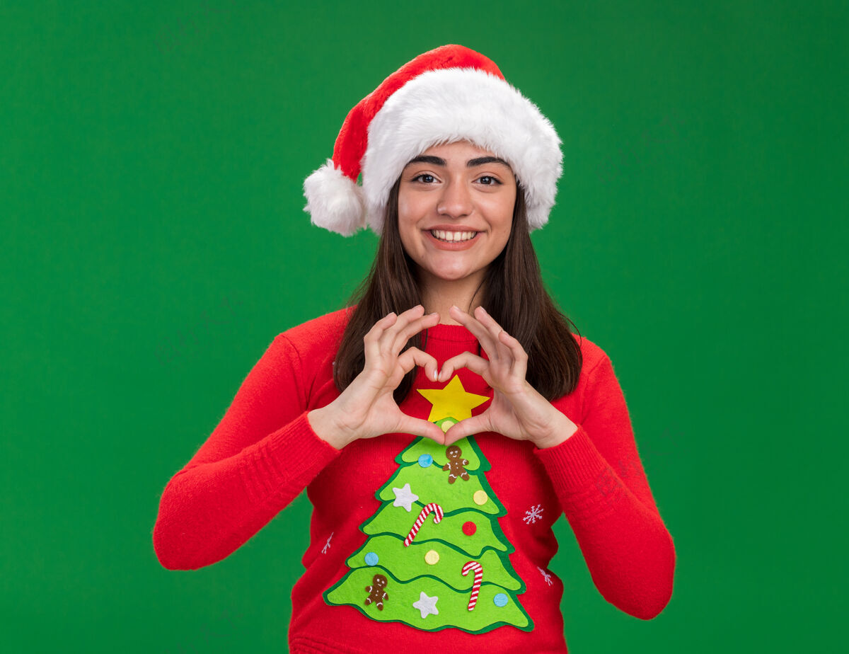 年轻微笑着的年轻白人女孩戴着圣诞帽 在绿色的背景上用复制的空间做着孤立的心形手势手势微笑心