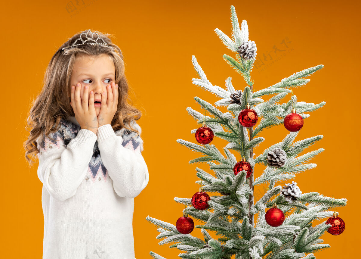 盖站在圣诞树旁的关心的小女孩戴着头饰 脖子上戴着花环 两手孤立在橙色的墙上戴小头饰