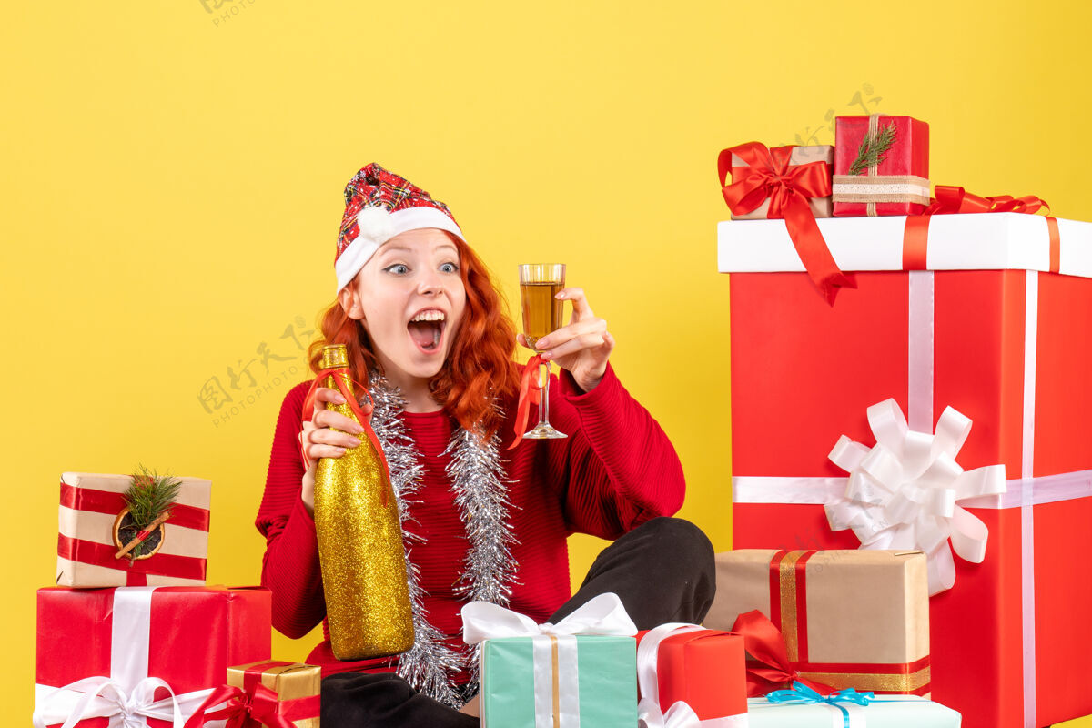 女性年轻女子坐在圣诞礼物周围 在黄色墙上用香槟庆祝的正面视图香槟前面年轻女性