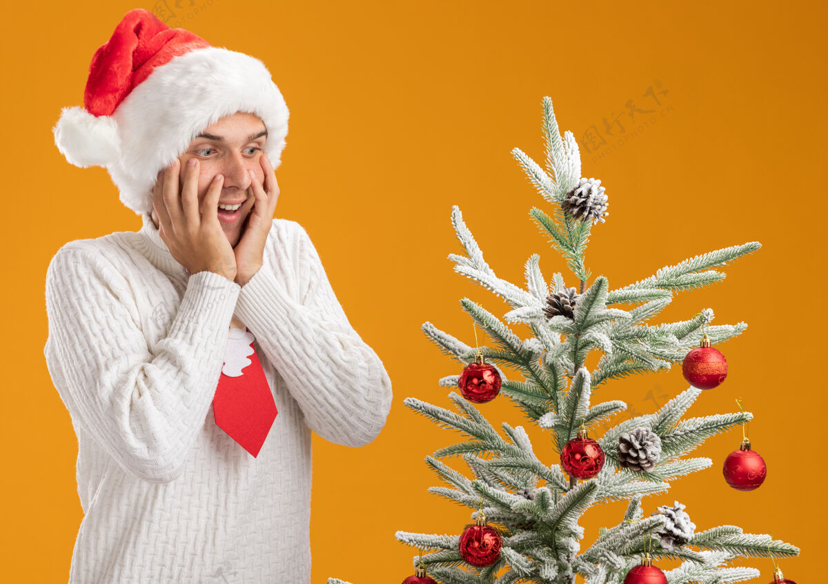 年轻的年轻帅哥戴着圣诞帽 打着圣诞老人的领带 站在装饰好的圣诞树旁 手放在脸上 孤零零地看着橙色的墙上靠近领带印象深刻