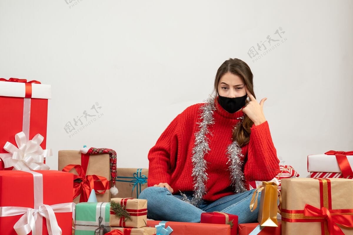 周围穿着红色毛衣 戴着黑色面具的年轻女孩把手指枪放在太阳穴里 围着白色的礼物坐着礼物放成人